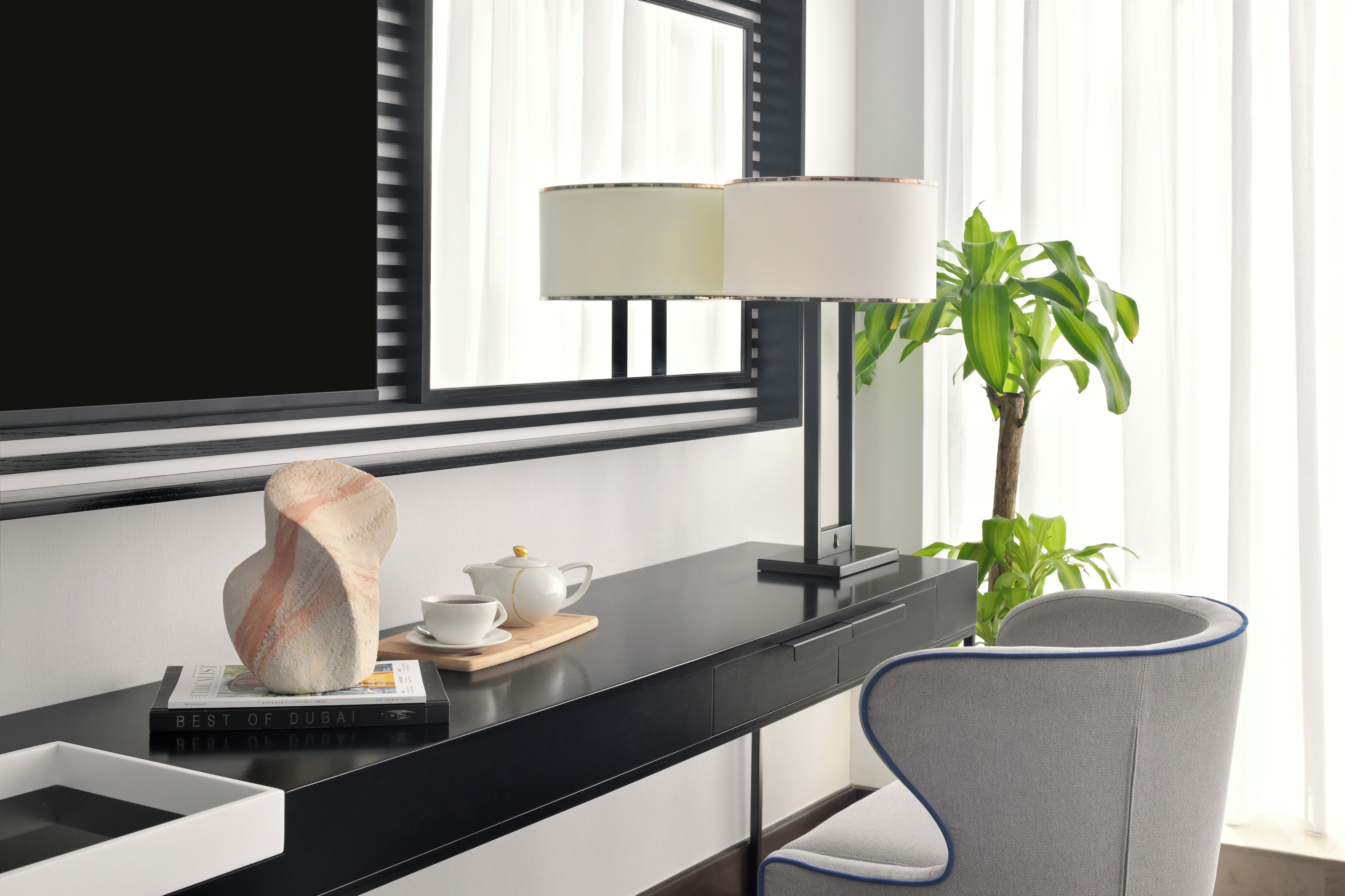 A modern hotel room with a black desk, a white lamp, a potted plant, a chair, and a large mirror.