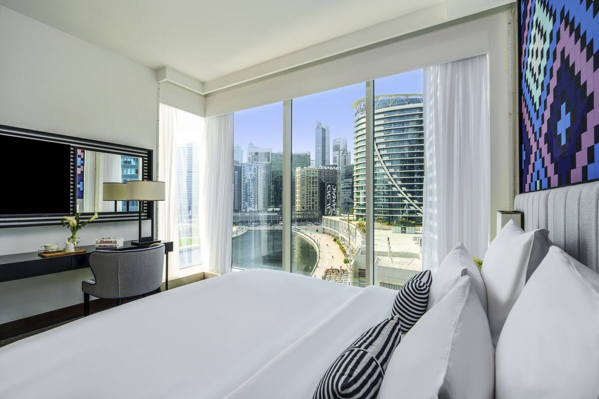 A hotel room with a large bed, a desk with a lamp, a chair, and a view of a cityscape through large windows.