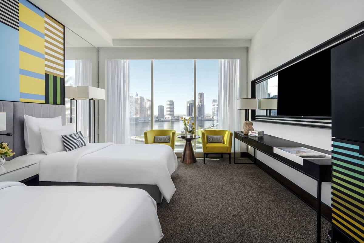 A hotel room with two beds, a desk, a chair, a TV, and a large window with a city view.