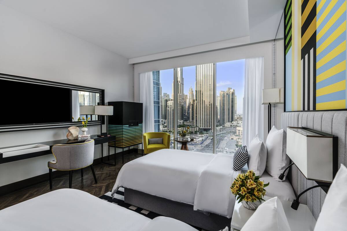 A modern hotel room with two beds, a TV, a desk, and a view of the city skyline.