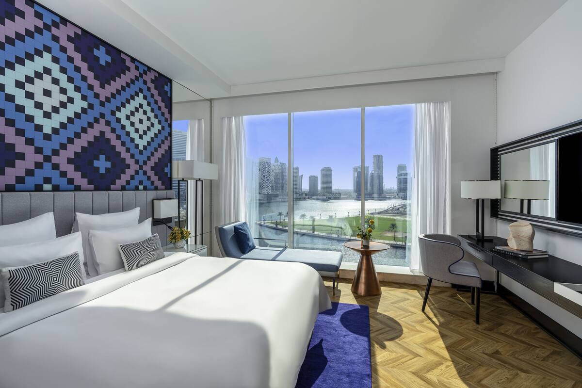 A modern hotel room with a large bed, a desk, a chair, and a view of the cityscape through large windows.