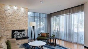 Un salon moderne avec un mur en pierre, une cheminée, un canapé, une table basse ronde et une vue dégagée sur l’extérieur grâce à de grandes fenêtres ornées de rideaux légers.