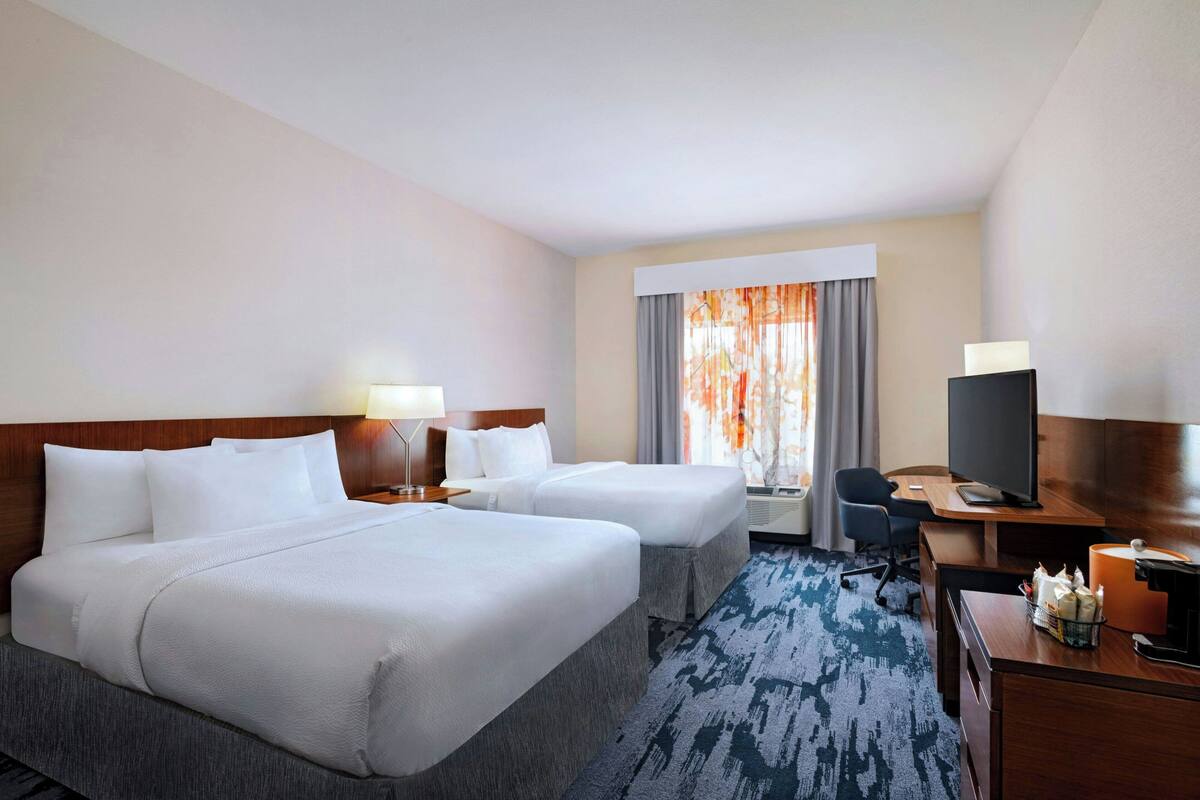 A hotel room with two beds, a desk, a TV, and a window with curtains.