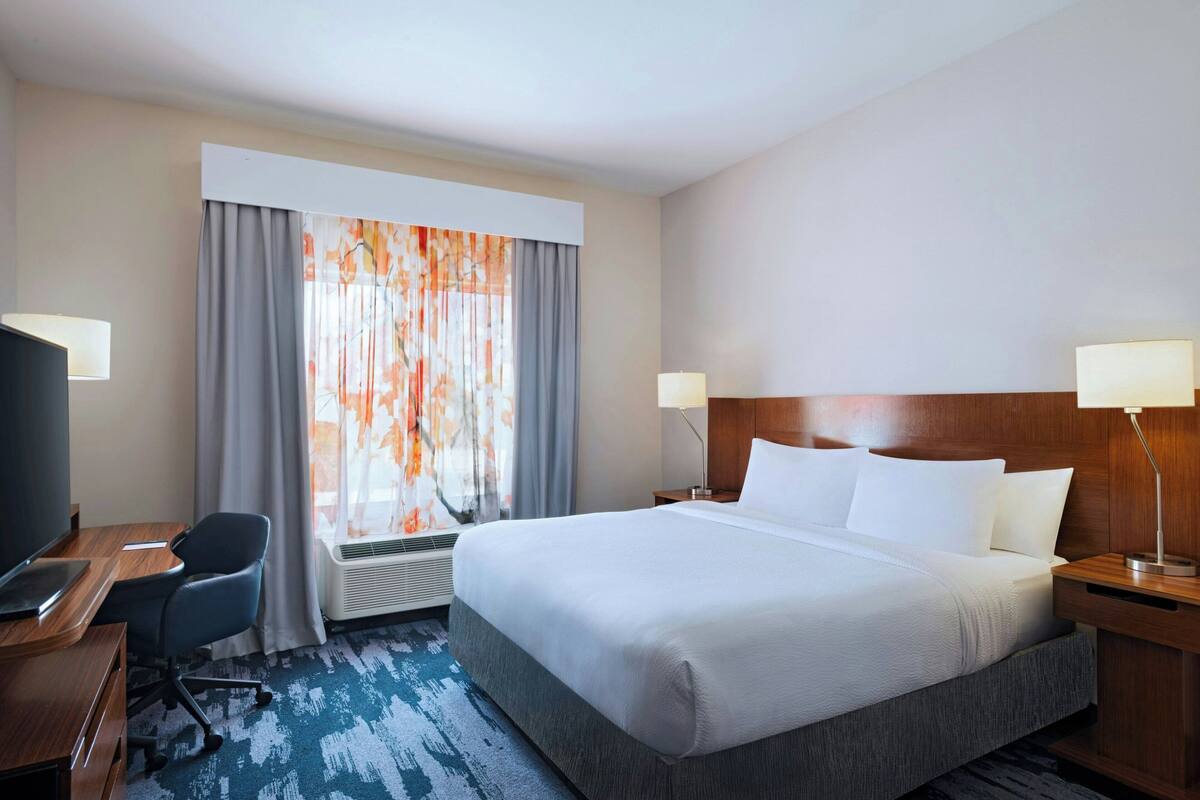 A hotel room with a large bed, a desk with a chair, a TV, and a window with curtains.