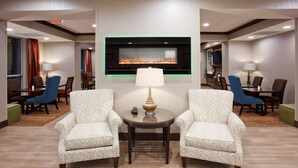 A hotel lobby with two armchairs facing a fireplace, a round table with a lamp, and a television mounted on the wall. - Hampton Inn Minneapolis Bloomington West (Bloomington)