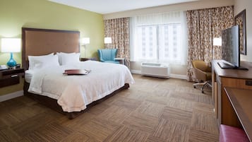 A hotel room with a large bed, a desk with a computer, a chair, a TV, and an air conditioning unit.