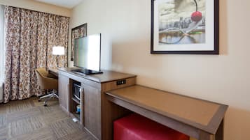 A hotel room with a desk, a television, a red bench, a patterned curtain, and a framed picture on the wall.