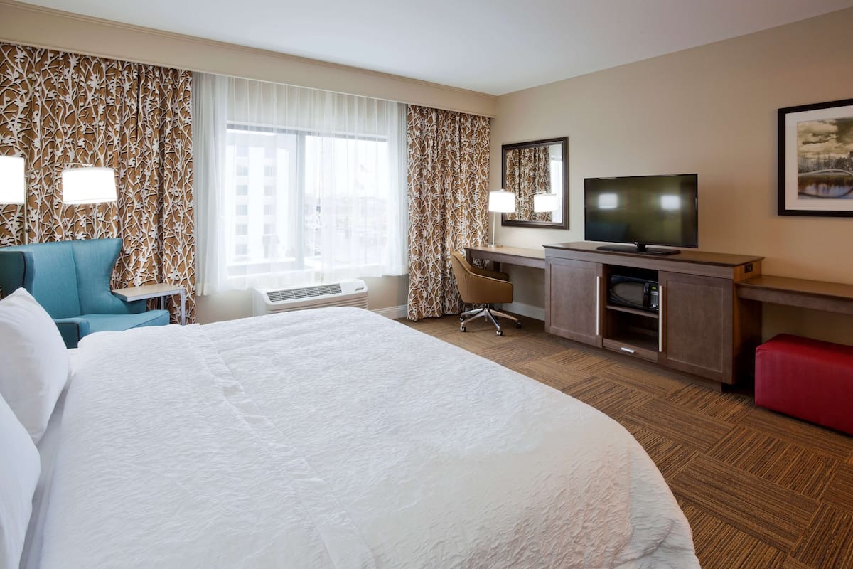 A hotel room with a large bed, a desk with a TV, a chair, and a red bench.