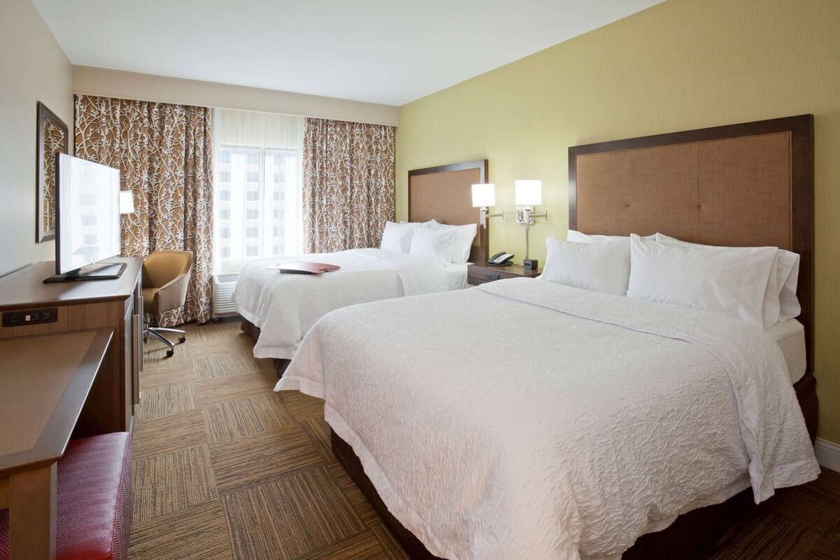 A hotel room with two beds, a desk, a chair, a television, and a window with curtains.