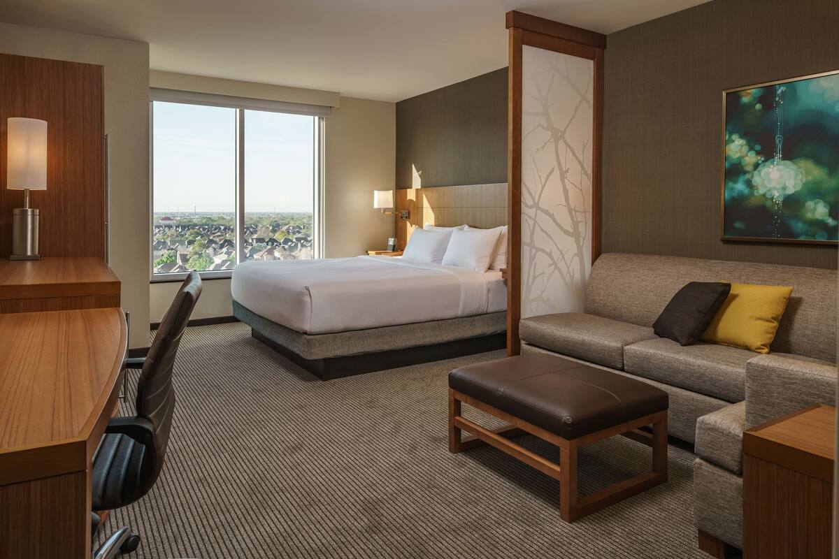 A hotel room with a large bed, a sofa, a desk, and a chair. There is a large window providing a view of the city.
