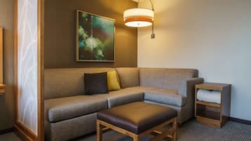 A hotel room with a sectional sofa, a wooden ottoman, a small side table, and a wall-mounted lamp.
