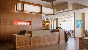 Zona de recepción de un hotel con un mostrador de madera, una máquina de café y un área de estar con sillas y una mesa.