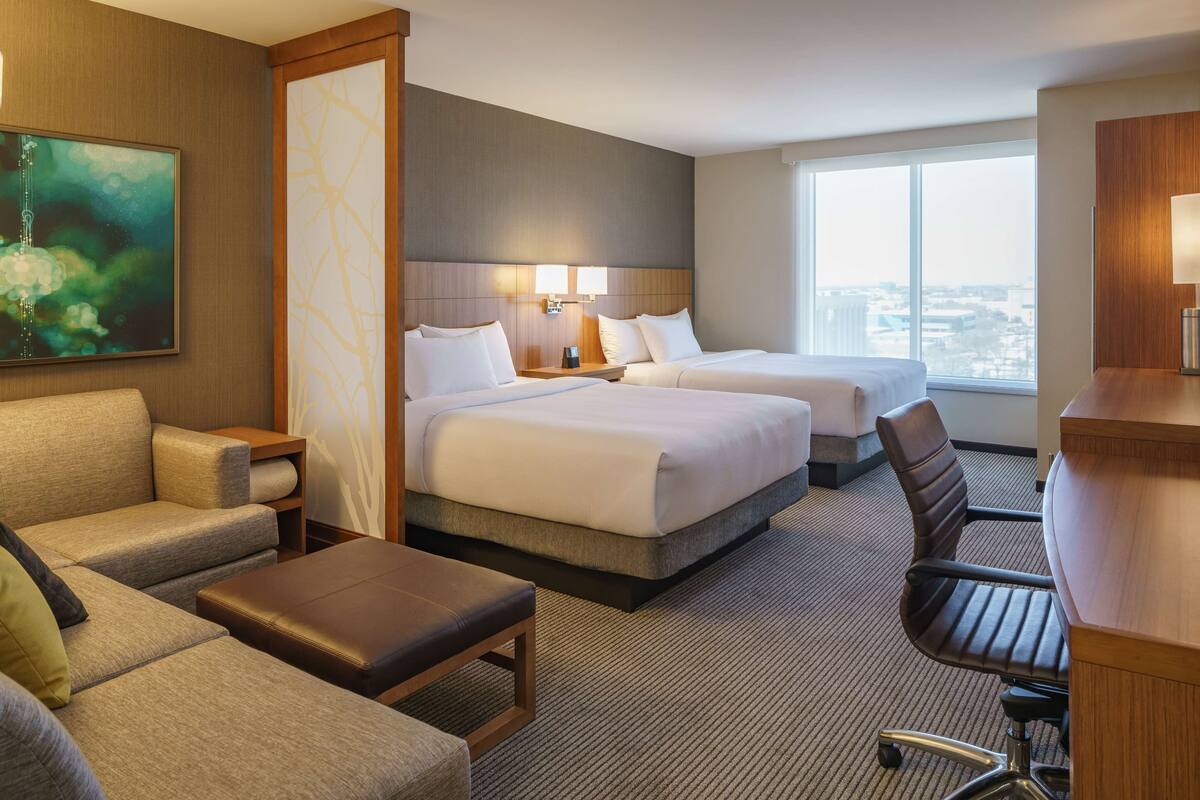 A hotel room with two beds, a sofa, a desk, and a large window offering a city view.