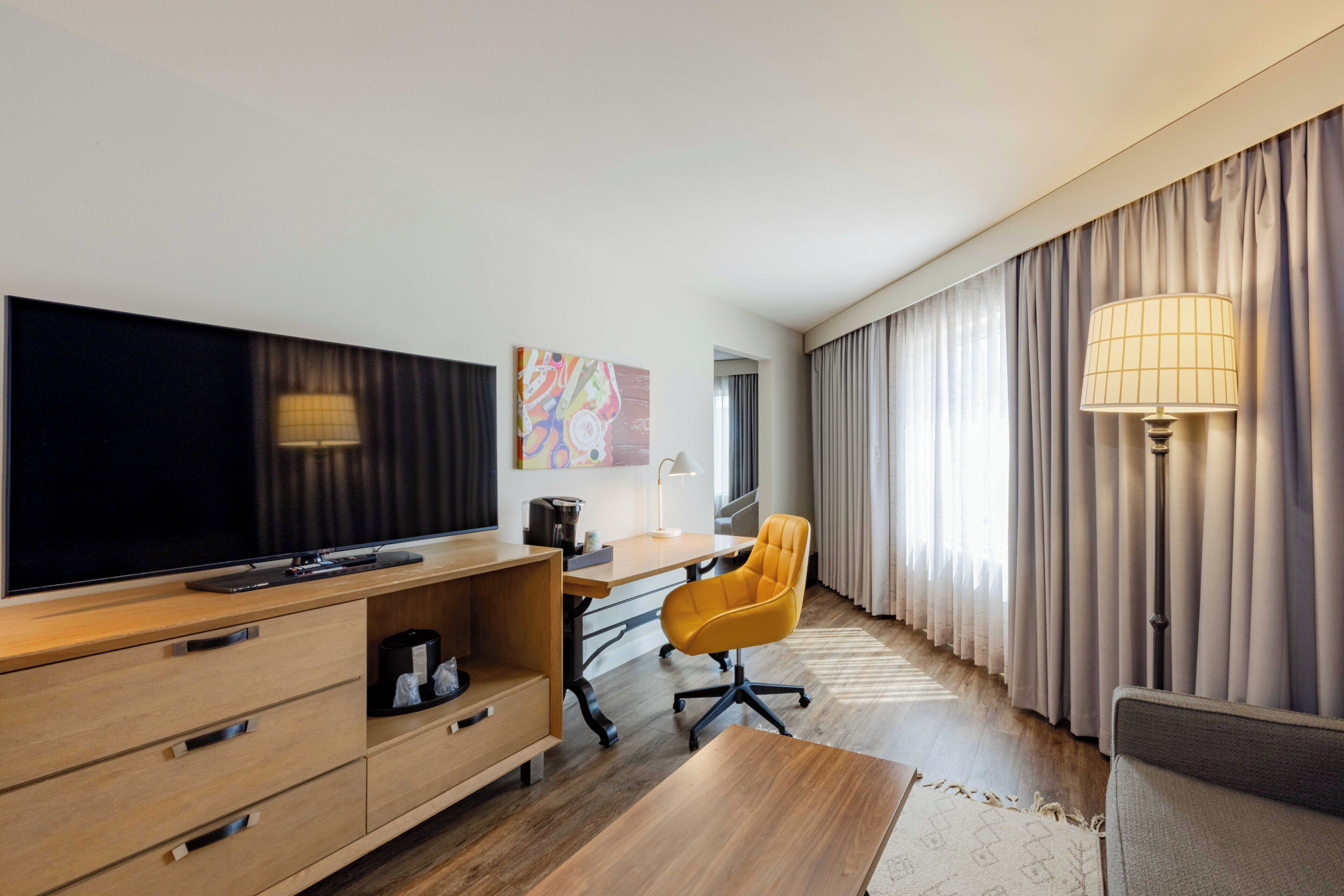 A modern hotel room with a flat-screen TV, wooden furniture, a desk, a yellow chair, and a large window with curtains.