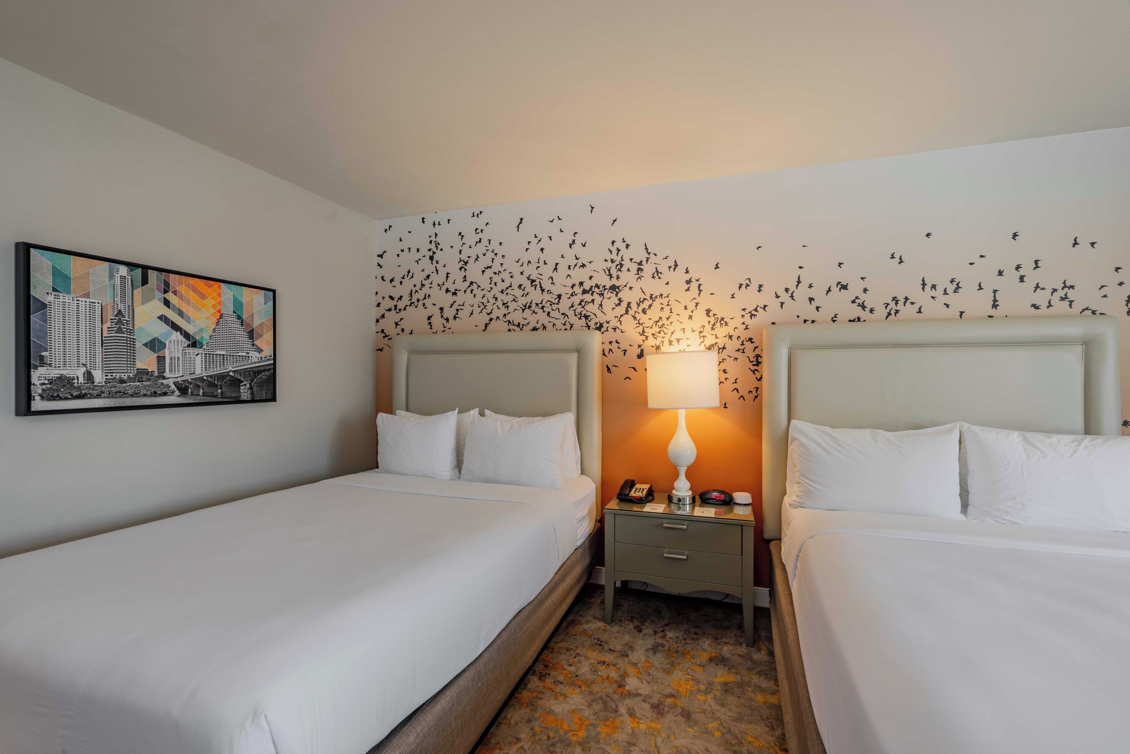 Une chambre d’hôtel avec deux lits simples, une table de chevet avec une lampe et une œuvre d’art accrochée au mur représentant un paysage urbain.