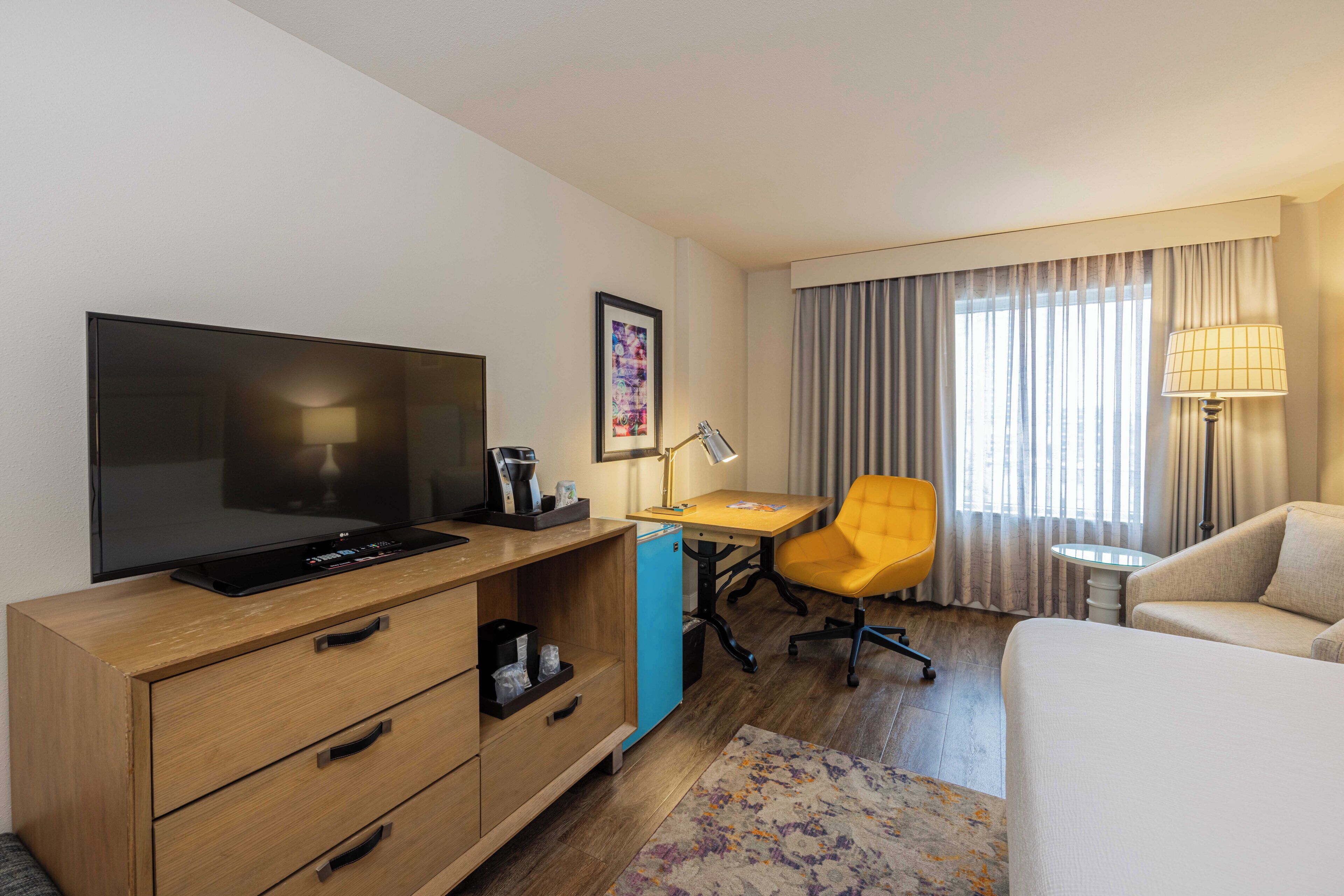 Habitación de hotel con televisión de pantalla plana, escritorio con lámpara, silla amarilla, sofá y cama.
