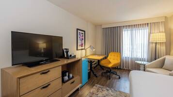 Habitación de hotel con televisión de pantalla plana, escritorio con lámpara, silla amarilla, sofá y cama.