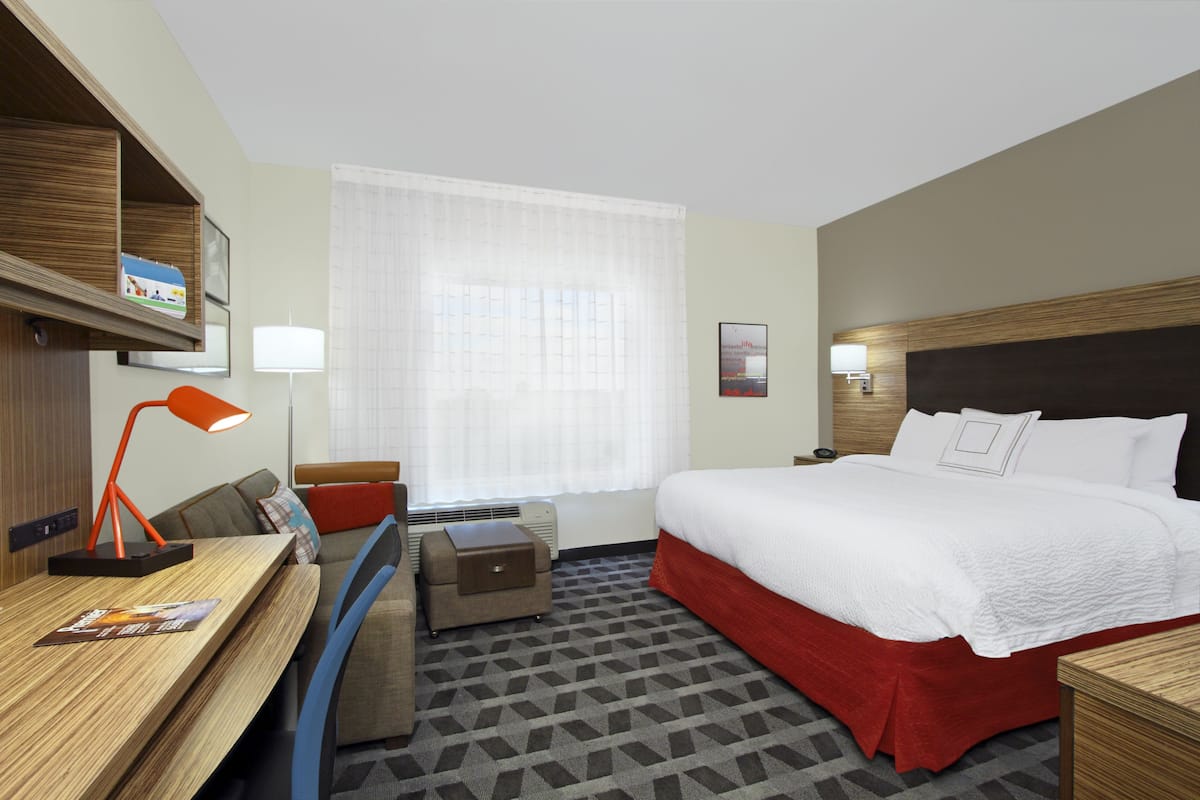 A hotel room with a bed, a desk, a chair, a small table, a lamp, a nightstand, and a window with curtains.