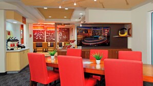 A modern hotel lobby with a large dining area featuring red chairs and a wooden table, a reception desk, and a wall-mounted television displaying a scenic view.