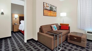 A hotel room with a sofa, a small table, a bedside table, a lamp, and a painting on the wall.