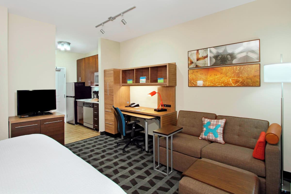 A hotel room with a bed, a desk with a chair, a sofa, a TV, a kitchenette, and artwork on the wall.