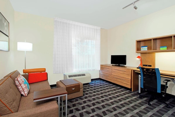 Habitación de hotel con sofá, una mesita, escritorio con computadora, televisor y ventana con cortinas.