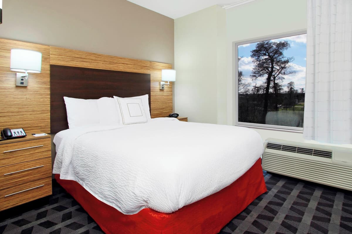 A hotel room with a large bed, wooden headboard, bedside lamps, a telephone, a window with a view of trees, and a radiator.