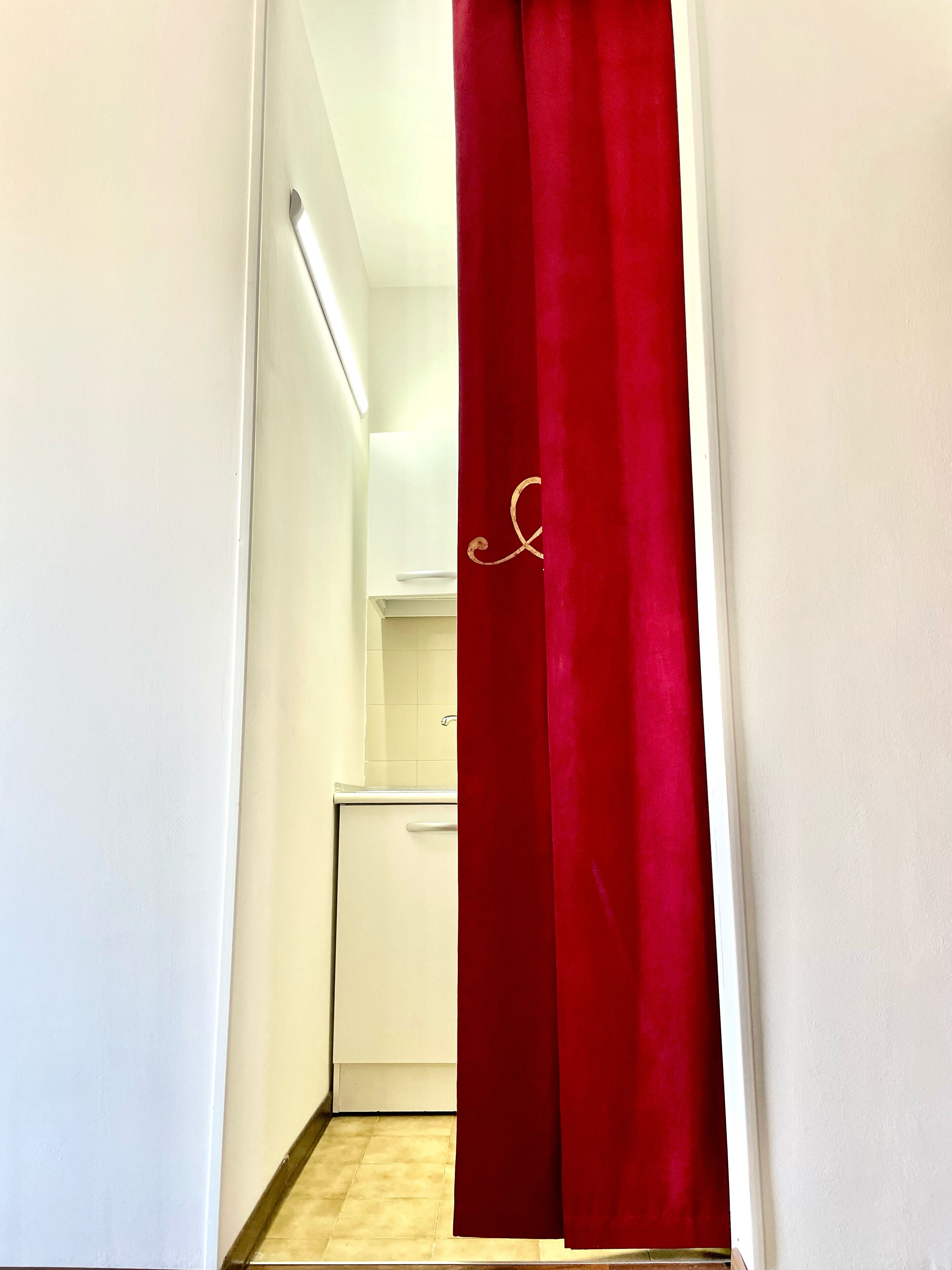 Un estrecho pasillo con una cortina roja que presenta un símbolo dorado, que conduce a una zona de cocina con armarios blancos y una encimera blanca.