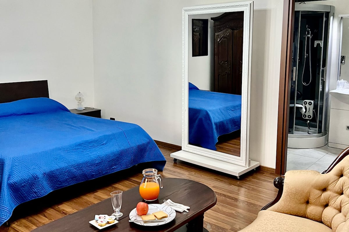 A hotel room with a bed, a chair, a table with breakfast, a mirror, and a shower area.