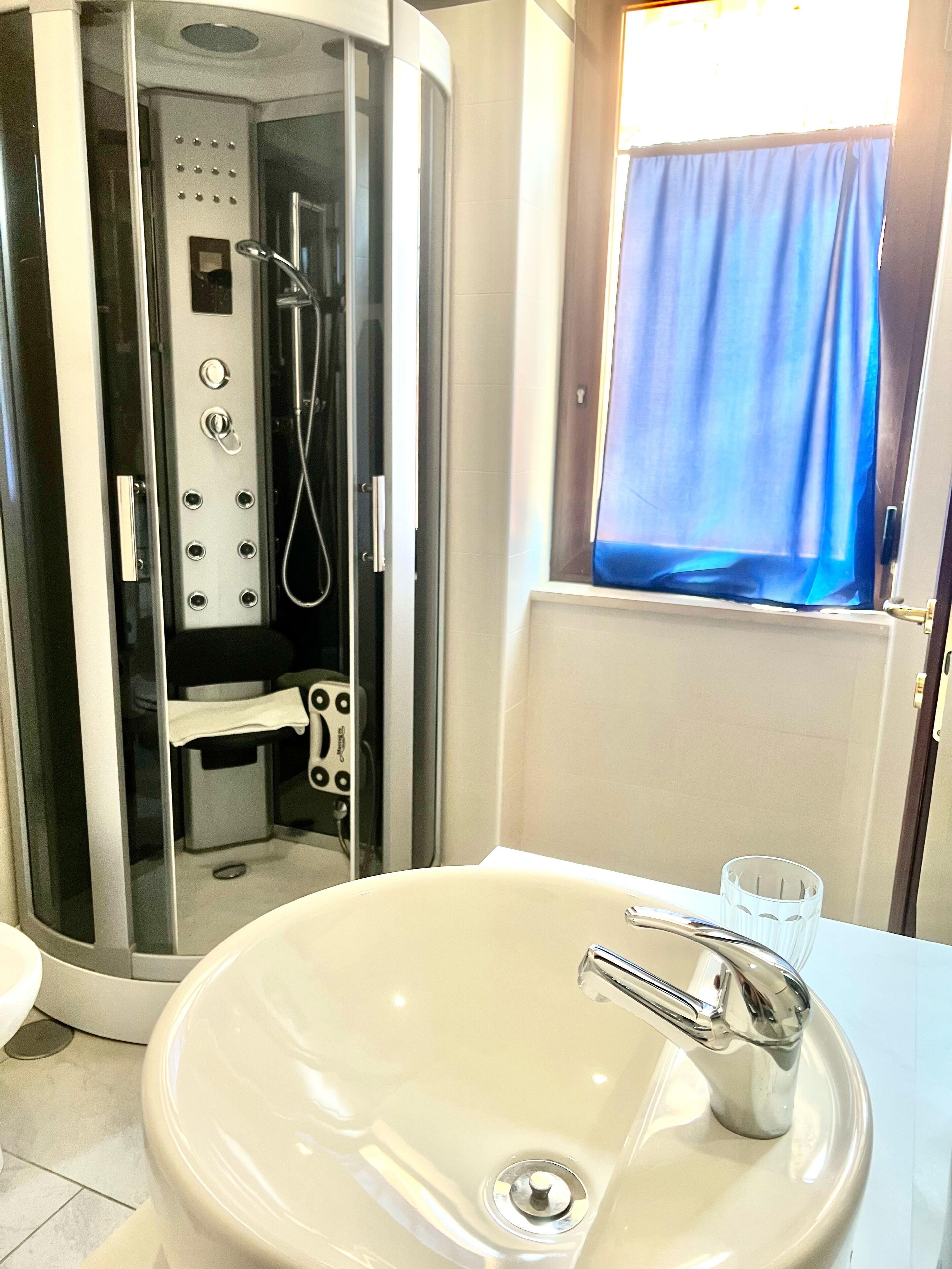 Une salle de bain moderne avec une douche en verre, un lavabo blanc et une fenêtre agrémentée d’un rideau bleu.