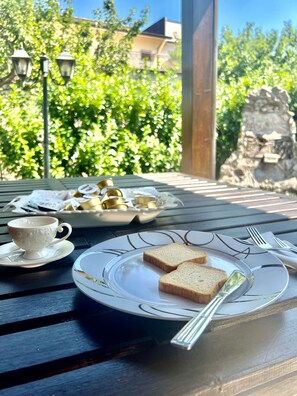 Una mesa servida con tostadas, una taza y una bandeja de pasteles, con una escultura de piedra y follaje al fondo.