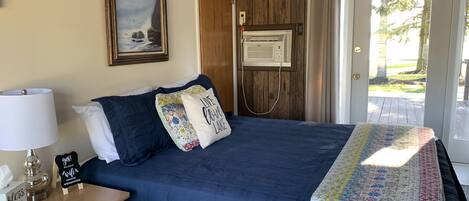 Un dormitorio con cama, mesita de noche, lámpara, cuadro enmarco, termostato y puerta que da al exterior.