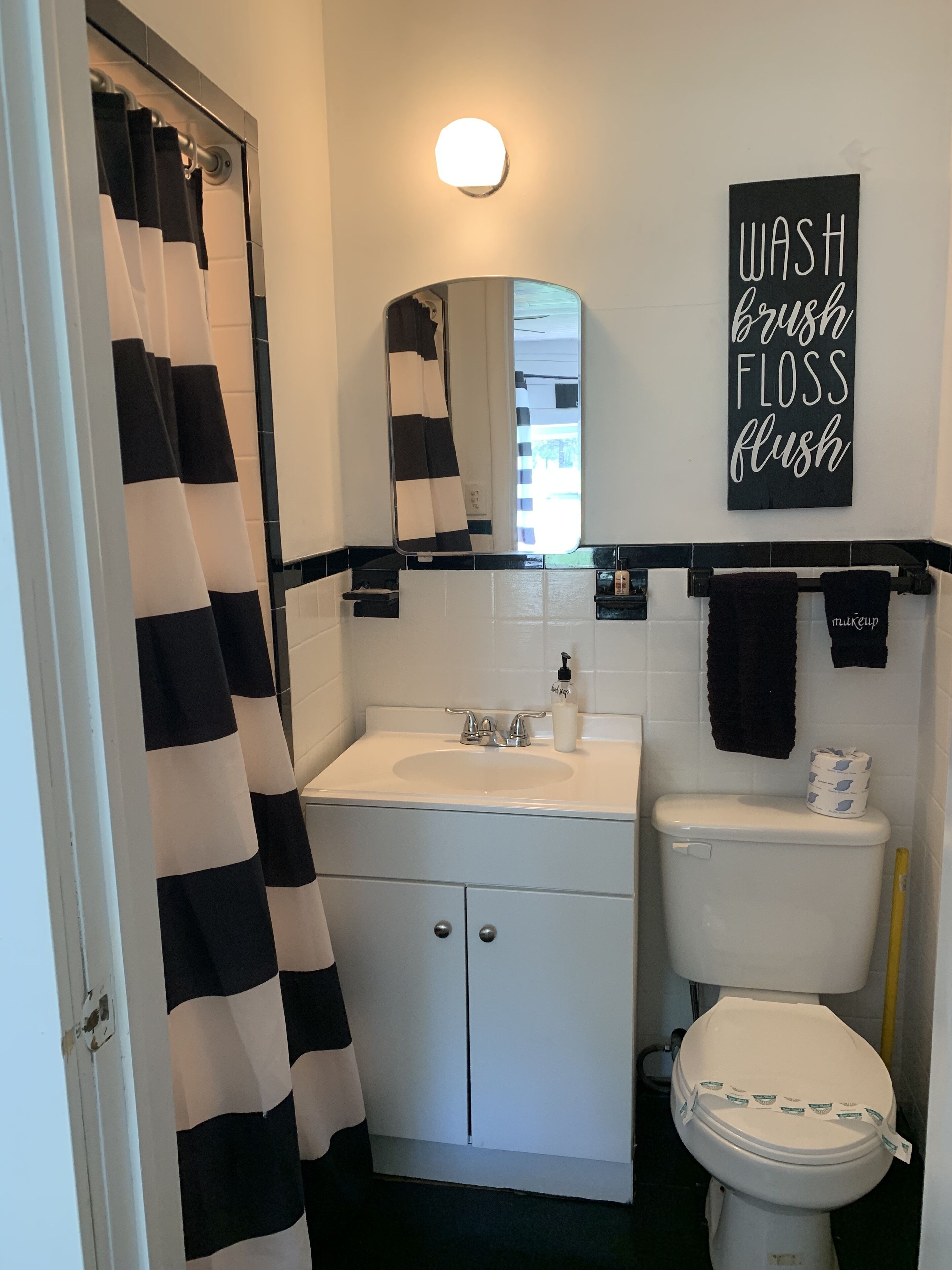 A bathroom with a white sink, black and white striped shower curtain, a toilet, and a wall-mounted sign that reads "WASH BRUSH FLOSS FLUSH".