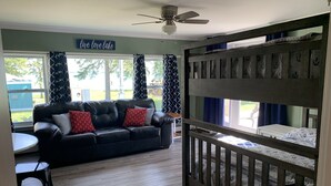 Un salon avec un canapé en cuir noir, un lit superposé en bois avec une literie ornée de motifs d’ancre, un ventilateur de plafond et un panneau indiquant : « Vivez, aimez, profitez du lac ».