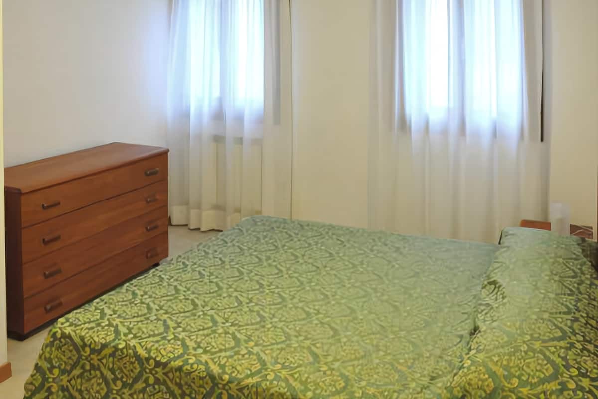 Een slaapkamer met een houten dressoir, een bed met een gestreept sprei en twee ramen met witte gordijnen.
