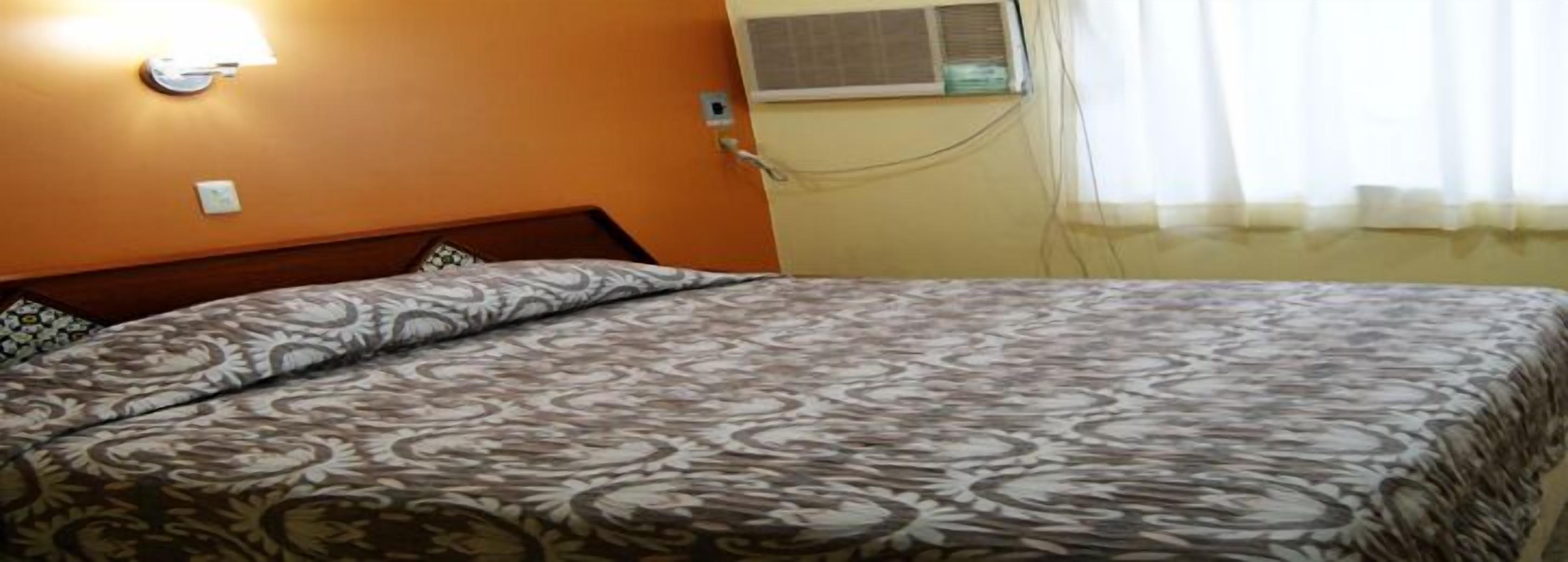Una cama con una colcha estampada, un equipo de aire acondicionado en la pared y una ventana con cortinas.