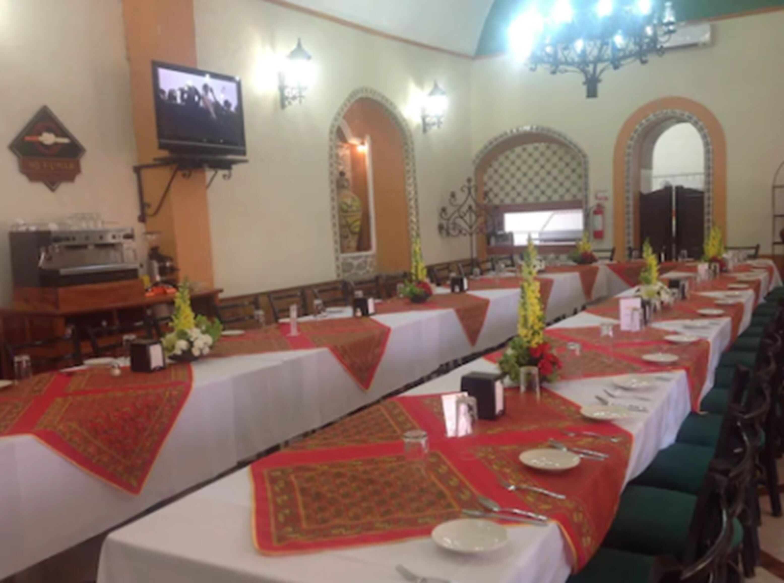 Uma longa mesa de jantar com toalhas vermelhas e centros de mesa, em um salão com janelas em arco e um lustre.