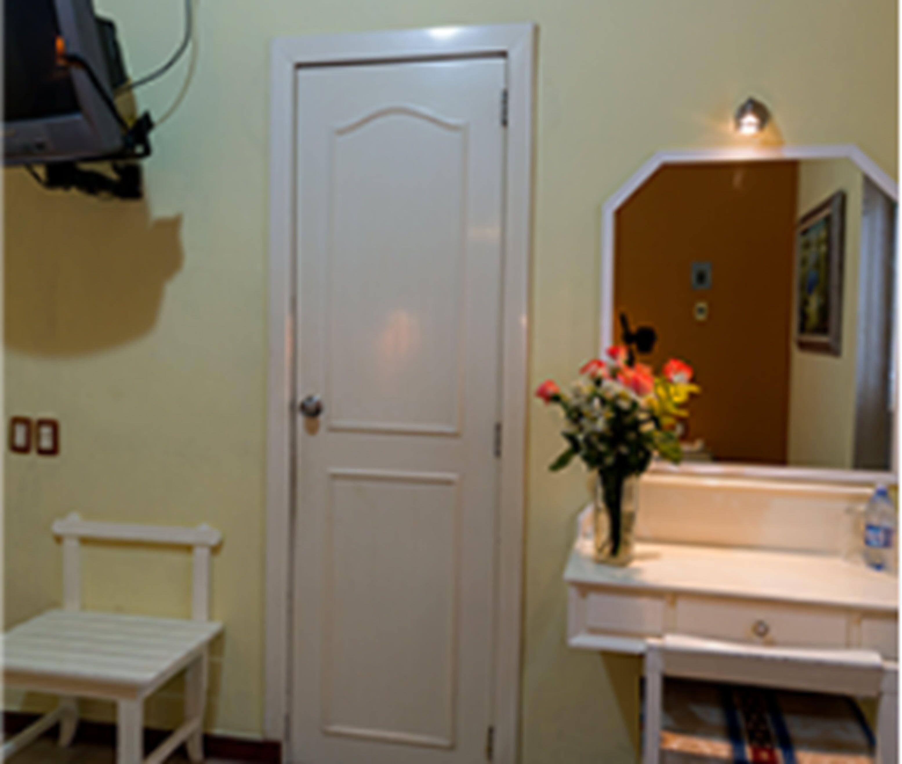 A room with a white door, a small white chair, a vanity with a mirror, and a vase with flowers.
