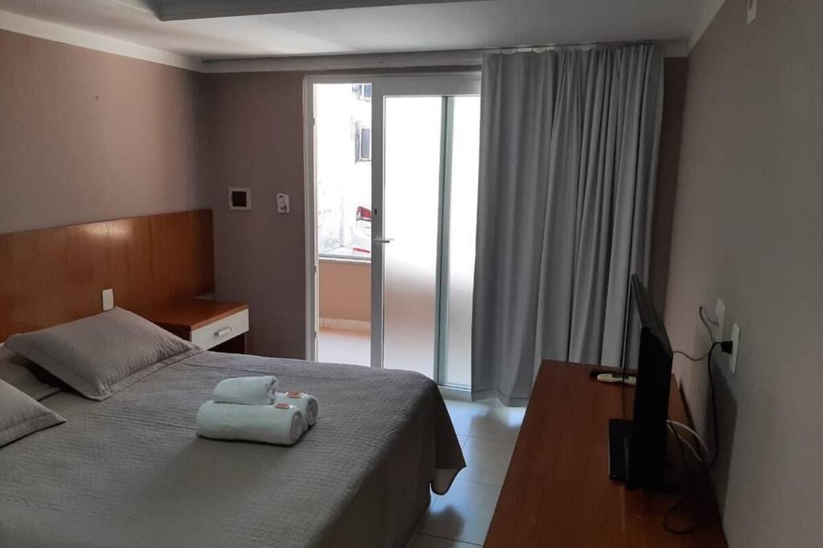 Quarto de hotel com uma cama, uma escrivaninha, uma televisão e uma porta de correr para uma varanda.