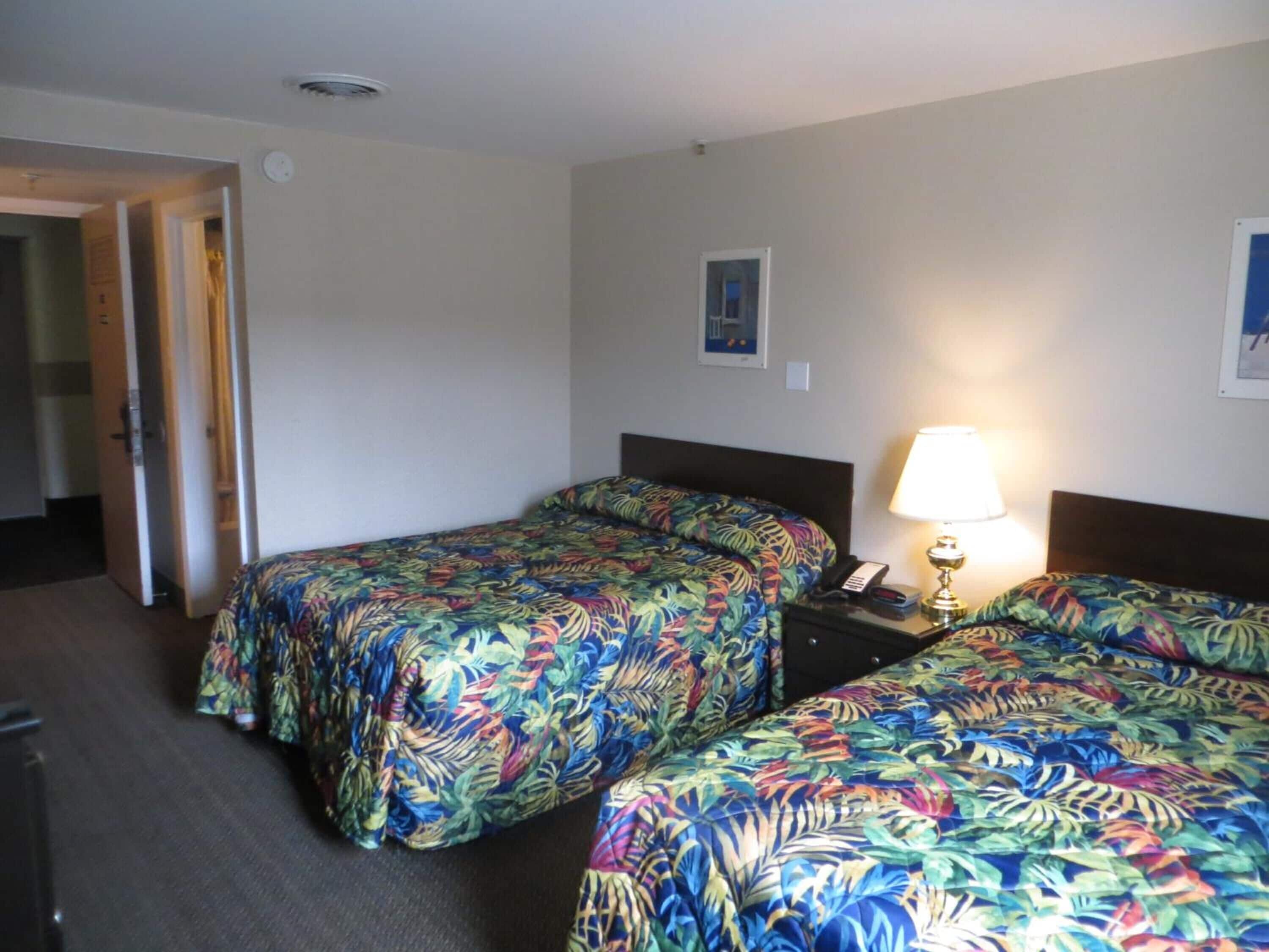 Habitación de hotel con dos camas, una mesita de noche con lámpara, y una puerta que da a otra habitación.