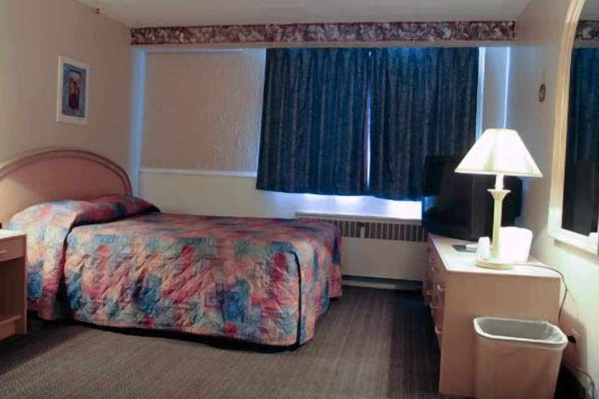 Hotellihuone, jossa on sänky, kirjoituspöytä lampun kanssa, tuoli ja ikkuna, jossa on verhot.