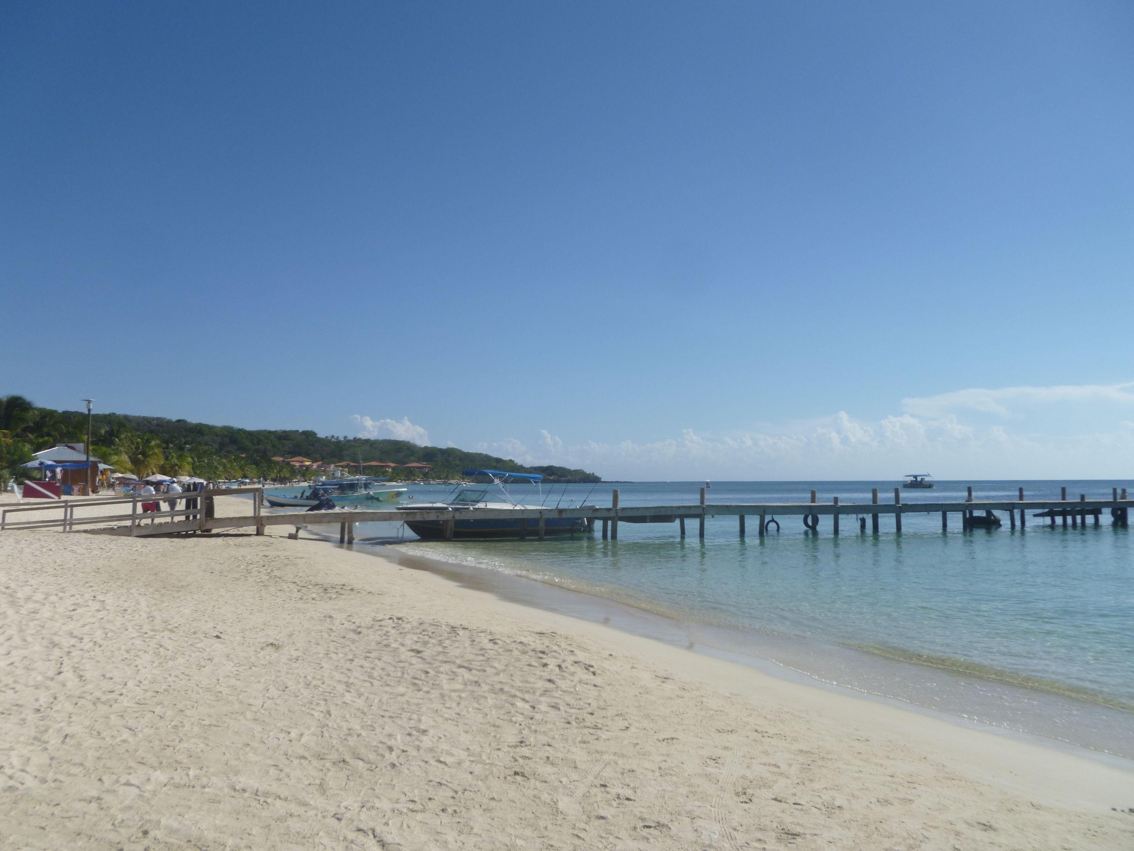 Une plage avec une jetée en bois s’avançant dans des eaux calmes, un rivage sablonneux et quelques bateaux ancrés à proximité.
