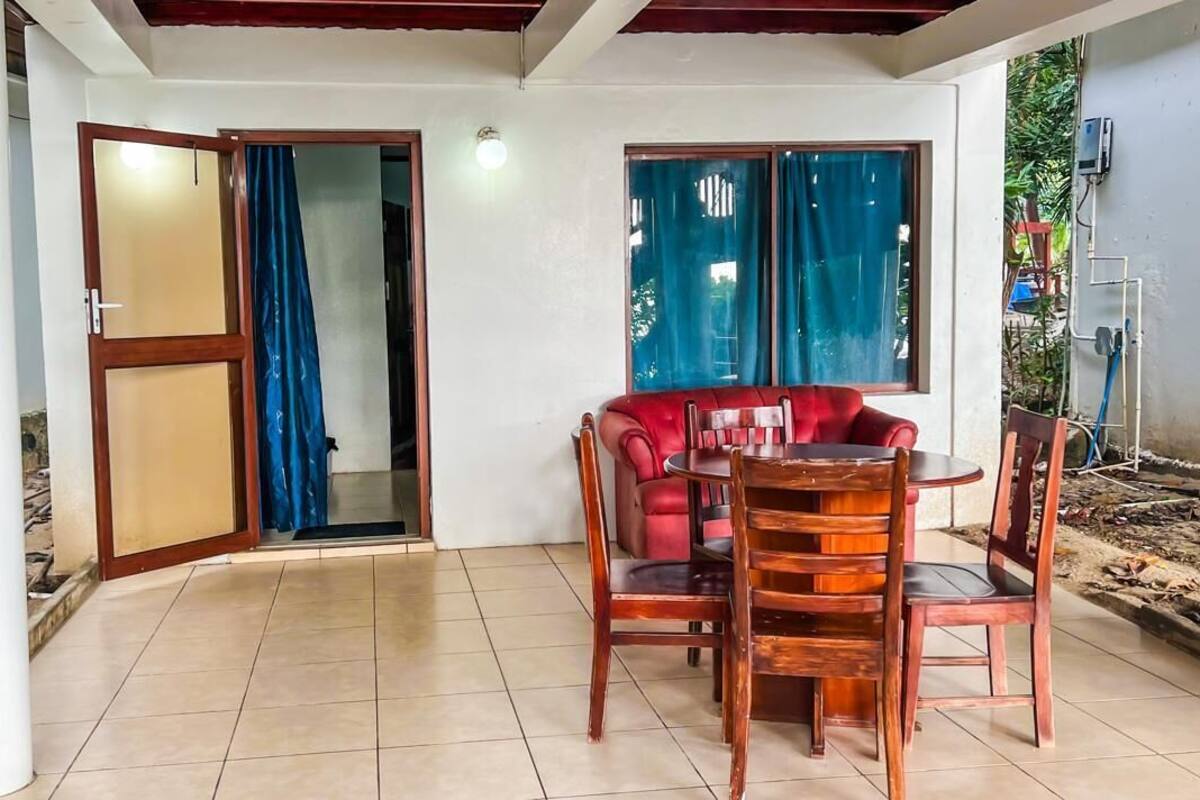 A covered patio area with a red sofa and wooden chairs, a tiled floor, and a sliding glass door leading to an interior space.