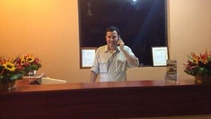 Een receptiebalie in een hotel, met een man aan de telefoon, foto's aan de muur en bloemstukken.