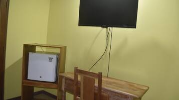 Una habitación con un escritorio de madera, un refrigerador miniatura blanco y un televisor de pantalla plana montado.
