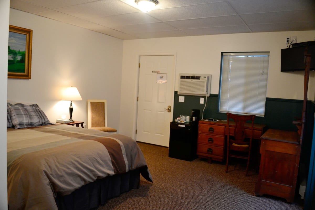 A hotel room with a bed, a desk, a chair, a dresser, a lamp, an air conditioning unit, and a door.