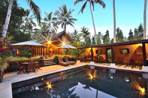 An outdoor pool area with lounge chairs and umbrellas, surrounded by palm trees and a building with a thatched roof.