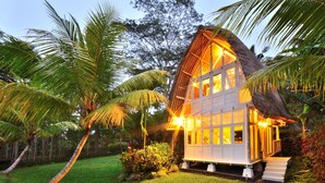 Une maison à toit de chaume, dotée de nombreuses fenêtres, entourée d’une végétation luxuriante et de palmiers.