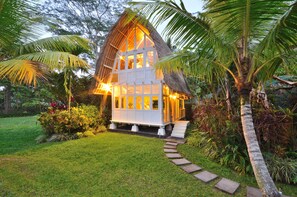 A thatched-roof house with a pathway leading to it, surrounded by lush greenery and palm trees.