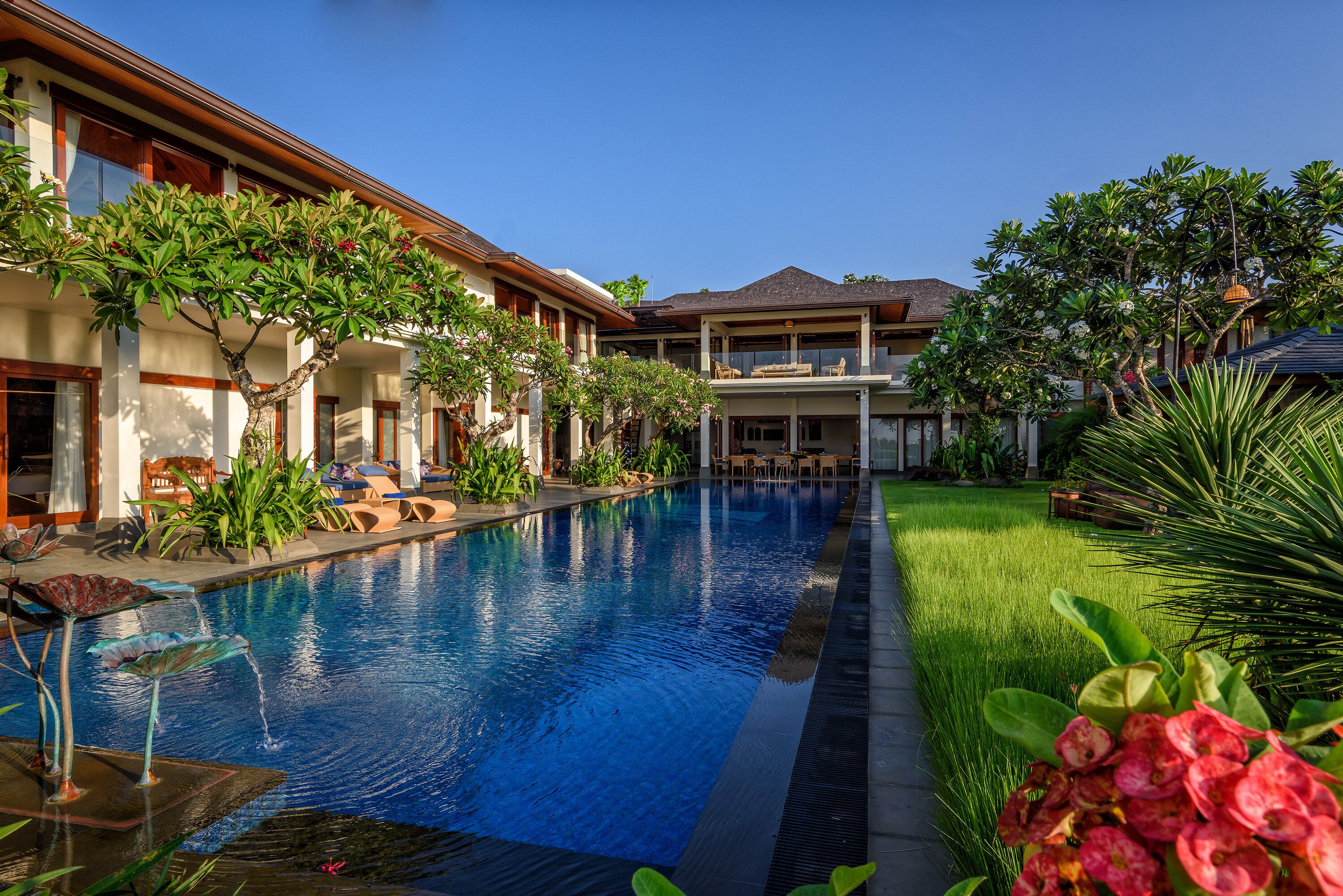 A swimming pool with a clear blue water surface, surrounded by lush greenery and a building with a balcony.